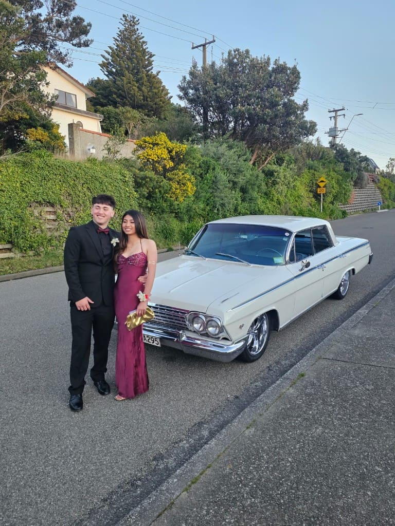 A couple dressed for a formal standing in front of the white Impala on a suburban road.