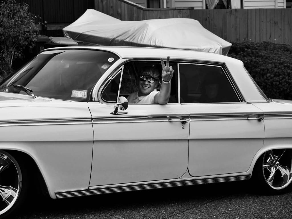 Liam McLean leaning from the driver's window of the white Impala, wearing a flat cap and sunglasses.